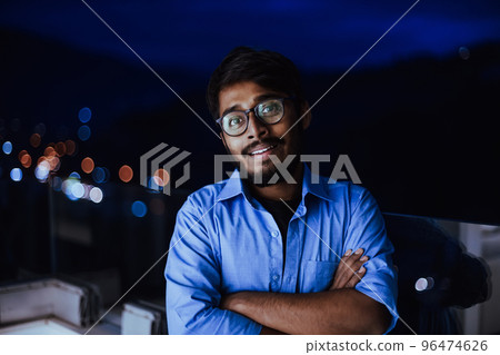 An Indian man with glasses and a blue shirt looks around the city at night. In the background of the night street of the city An Indian man with glasses and a blue shirt looks around the city at night. In the background of the night street of the city 96474626
