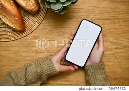 A smartphone white screen mockup is in a woman's hand over wooden tabletop. top view A smartphone white screen mockup is in a woman's hand over wooden tabletop. top view 96474882