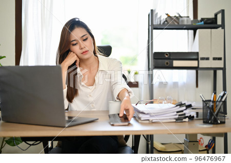 Bored and tired Asian businesswoman at her office desk, about to use her smartphone. 96476967