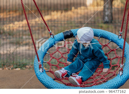 Cute Little boy with down syndrome in a funny hat walks in the playground, swinging on a swing 96477162