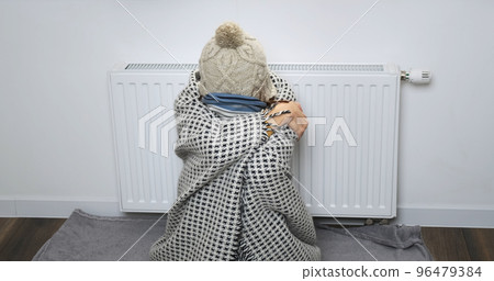 Funny woman in warm clothes freezing at home. Girl puts on hat in white room, pulls scarf over her nose, freezes indoors. Woman backdrop of central heating battery trying to warm herself Funny woman in warm clothes freezing at home. Girl puts on hat in white room, pulls scarf over her nose, freezes indoors. Woman backdrop of central heating battery trying to warm herself 96479384