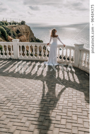 Romantic bride, blond girl in white wedding dress with open shoulders posing on open terrace with backdrop of the sea and rocks. Stylish young woman standing on terrace and looking on ocean at sunset Romantic bride, blond girl in white wedding dress with open shoulders posing on open terrace with backdrop of the sea and rocks. Stylish young woman standing on terrace and looking on ocean at sunset 96484375