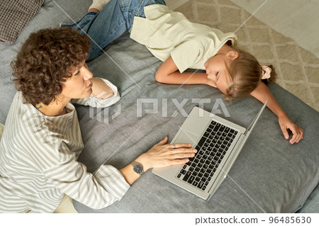 High angle view of mother talking to her little daughter while they relaxing on bed and using laptop High angle view of mother talking to her little daughter while they relaxing on bed and using laptop 96485630