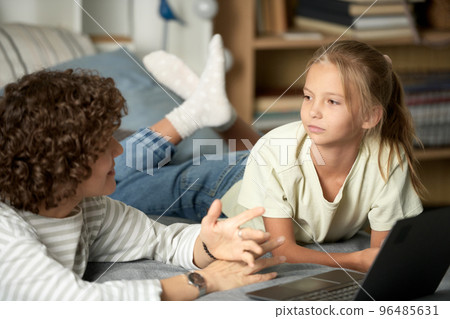 Little girl listening to her mom while she explaining her something, they lying on bed and using laptop Little girl listening to her mom while she explaining her something, they lying on bed and using laptop 96485631