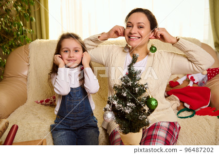 Cheerful mother and daughter have fun together, put shiny toy balls as earrings, decorate a Christmas tree at home. New Year's preparations. Magical festive atmosphere of the upcoming winter holidays Cheerful mother and daughter have fun together, put shiny toy balls as earrings, decorate a Christmas tree at home. New Year's preparations. Magical festive atmosphere of the upcoming winter holidays 96487202