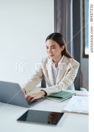 Portrait of a young Asian woman showing a smiling face as she using computer and financial documents on her desk in the early morning hours. 96487356