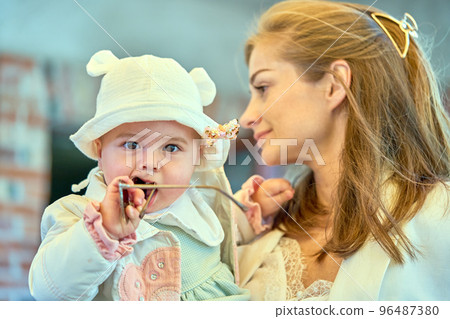A selective focus shot of a cute baby in her mother's arms taking sunglasses to her mouth 96487380