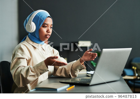 Young confident Muslim woman in headphones over blue hijab 96487813