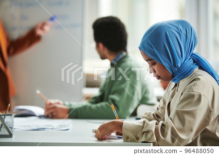 Side view of young serious Muslim woman in blue hijab making notes 96488050