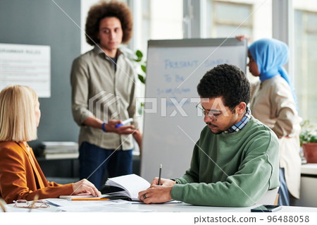 Young man making notes in copybook while sitting by desk at lesson Young man making notes in copybook while sitting by desk at lesson 96488058