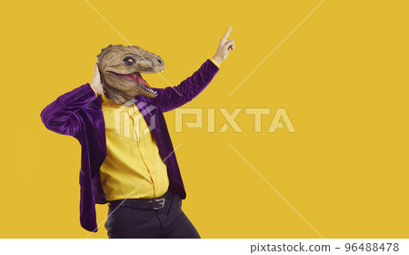 Eccentric showman in dinosaur mask and velvet jacket pointing index finger on yellow background. 96488478