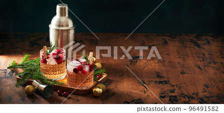 Alcoholic cocktail or non-alcoholic cocktail with vodka and cranberries with ice, shaker, jigger and bar spoon, fir branches and glowing garland for christmas on wooden background Alcoholic cocktail or non-alcoholic cocktail with vodka and cranberries with ice, shaker, jigger and bar spoon, fir branches and glowing garland for christmas on wooden background 96491582