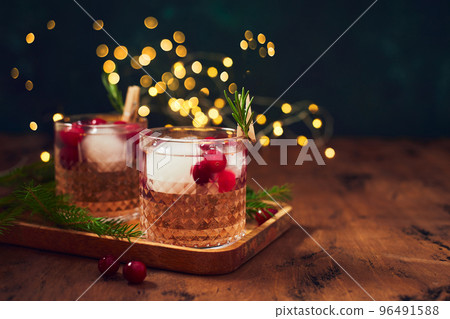 Alcoholic cocktail or non-alcoholic cocktail with vodka and cranberries with ice with fir branches and glowing garland for christmas on wooden background Alcoholic cocktail or non-alcoholic cocktail with vodka and cranberries with ice with fir branches and glowing garland for christmas on wooden background 96491588
