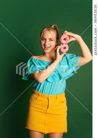 Emotional young beautiful girl, student in summer outfit is goofing around with glazed donuts isolated over green background. Positive emotions, happiness, hobbies and joy Emotional young beautiful girl, student in summer outfit is goofing around with glazed donuts isolated over green background. Positive emotions, happiness, hobbies and joy 96493636