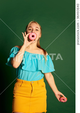 Happy young beautiful girl, student in summer outfit is goofing around with glazed donuts isolated over green background. Positive emotions, happiness, hobbies and joy Happy young beautiful girl, student in summer outfit is goofing around with glazed donuts isolated over green background. Positive emotions, happiness, hobbies and joy 96493640