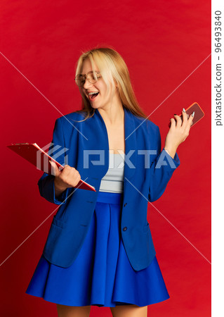 Positive young business woman or happy student wearing blue suit posing over bright red background. Startup, motivation, career, emotions, achievement concept Positive young business woman or happy student wearing blue suit posing over bright red background. Startup, motivation, career, emotions, achievement concept 96493840