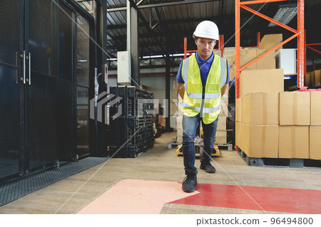 Asian male warehouse worker pulling a pallet truck. Worker working with hand pallet truck unloading cargo boxes on pallet at the warehouse. 96494800