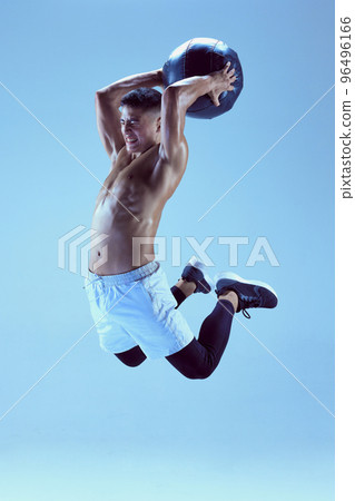 Portrait of young muscular man training with med ball, jumping isolated over blue background in neon light 96496166