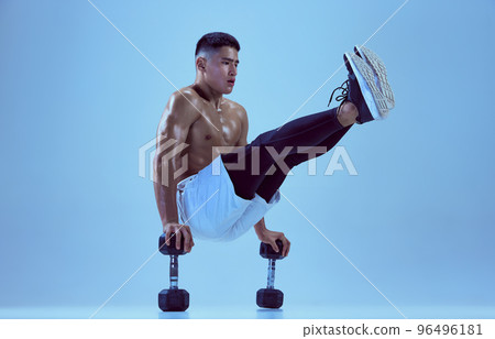 Portrait of young muscular man training, standing on dumbbells and lifting legs isolated over blue background in neon light Portrait of young muscular man training, standing on dumbbells and lifting legs isolated over blue background in neon light 96496181