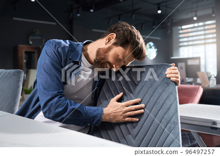 Romantic bearded man in casual outfit touching soft pattern, while leaning to chair indoors. Smiling brunet male with closed eyes hugging grey chair back, while sitting at table. Concept of emotions. Romantic bearded man in casual outfit touching soft pattern, while leaning to chair indoors. Smiling brunet male with closed eyes hugging grey chair back, while sitting at table. Concept of emotions. 96497257