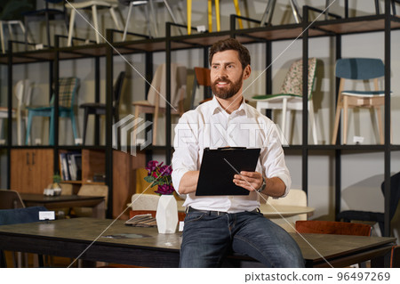 Happy bearded manager leaning on table, while working in furniture store. Portrait of pensive seller making notes, looking into distance, against rack with various chairs on background. Work concept. Happy bearded manager leaning on table, while working in furniture store. Portrait of pensive seller making notes, looking into distance, against rack with various chairs on background. Work concept. 96497269