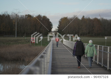 Male in orange hoodie running through national park across wooden bridge in wilderness in the cold fall weather. Sportsman does a fitness workout jogging on a timber footbridge in the countryside. Male in orange hoodie running through national park across wooden bridge in wilderness in the cold fall weather. Sportsman does a fitness workout jogging on a timber footbridge in the countryside. 96497328