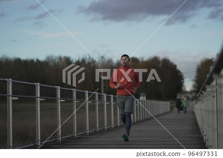 Cross country jogging in countryside. Man in orange sweatshirt running across wooden bridge in national park. Runner running over a wildlife trail wooden footbridge. Trail run in cold fall weather.  96497331