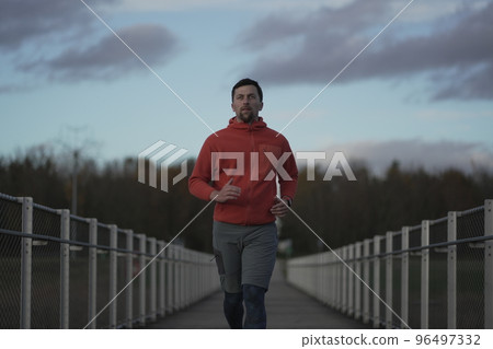Male in orange hoodie running through national park across wooden bridge in wilderness in the cold fall weather. Sportsman does a fitness workout jogging on a timber footbridge in the countryside.  96497332