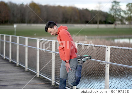 Athlete warming up before an outdoor cardio workout. A jogger warms up his muscles for a jog in nature on a wooden bridge in the cold fall weather. Healthy lifestyle. Rural running. Trail runs.  96497338