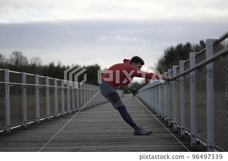 Man in orange and warm clothes warming up before jogging outdoors in cold autumn weather on wooden bridge. Runner stretching muscles leg and feet and preparing to run outdoors. Cross country running. Man in orange and warm clothes warming up before jogging outdoors in cold autumn weather on wooden bridge. Runner stretching muscles leg and feet and preparing to run outdoors. Cross country running. 96497339