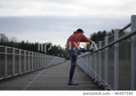 Man in orange and warm clothes warming up before jogging outdoors in cold autumn weather on wooden bridge. Runner stretching muscles leg and feet and preparing to run outdoors. Cross country running. Man in orange and warm clothes warming up before jogging outdoors in cold autumn weather on wooden bridge. Runner stretching muscles leg and feet and preparing to run outdoors. Cross country running. 96497340