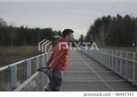 Man in orange and warm clothes warming up before jogging outdoors in cold autumn weather on wooden bridge. Runner stretching muscles leg and feet and preparing to run outdoors. Cross country running.  96497345