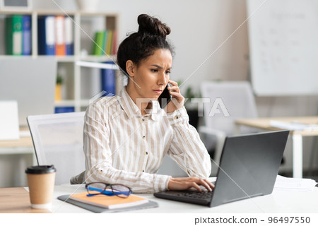 Serious businesswoman talking on smartphone and typing on laptop keyboard, sitting at workplace in office interior Serious businesswoman talking on smartphone and typing on laptop keyboard, sitting at workplace in office interior 96497550