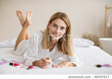 Attractive middle aged lady in solky bathrobe lying on bed and making manicure in bedroom interior, free space Attractive middle aged lady in solky bathrobe lying on bed and making manicure in bedroom interior, free space 96497617