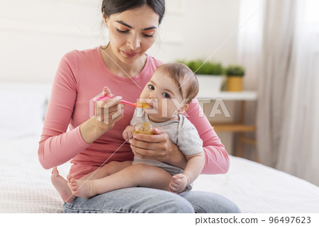 Adorable little baby girl eating from spoon, young caring mother feeding fer cute infant with mash fruit puree Adorable little baby girl eating from spoon, young caring mother feeding fer cute infant with mash fruit puree 96497623