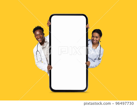Cheerful black young man and lady doctors in white coats peeking out from big phone with blank screen 96497730