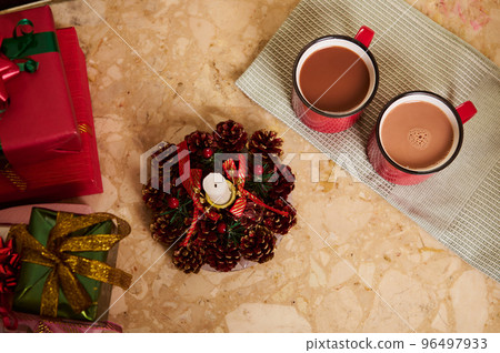 View from above of two red cups with hot cocoa or chocolate drink on a green napkin, next to a decorative Christmas lit candle and beautiful presents on marble table. New Year atmosphere at home. View from above of two red cups with hot cocoa or chocolate drink on a green napkin, next to a decorative Christmas lit candle and beautiful presents on marble table. New Year atmosphere at home. 96497933