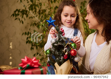 Mother and daughter decorating an eco Christmas tree with beautiful shiny bauble and Xmas toys. Christmastime. New Year's preparations and celebration. Enjoying happy winter holidays in family at home Mother and daughter decorating an eco Christmas tree with beautiful shiny bauble and Xmas toys. Christmastime. New Year's preparations and celebration. Enjoying happy winter holidays in family at home 96497942