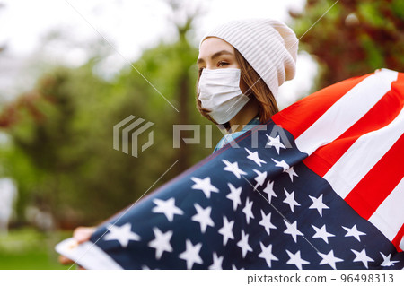 Girl in protective sterile medical mask and with american flag in hand. Quarantine city. Covid -19. Girl in protective sterile medical mask and with american flag in hand. Quarantine city. Covid -19. 96498313