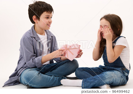 Handsome Caucasian teenage boy, loving caring brother greeting her little sister, who covers her eyes with hands, giving her a cute present in heart-shaped gift box. Saint Valentine's Day. Women's Day 96499462