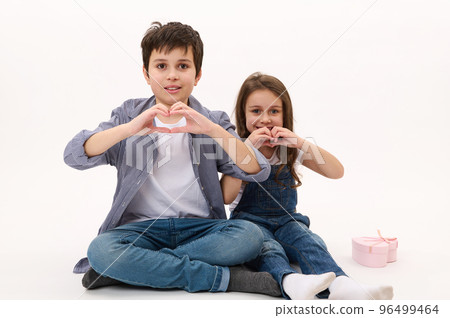 Adorable Caucasian children, boy and girl wearing casual shirt and denim jeans, gesturing showing a heart from fingers, smiling looking at camera, isolated over white background. Saint Valentines Day 96499464