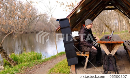 Man work on laptop charging from portable solar panel utdoor in alclove near the river. Clean energy for using in camping. Work with gadgets outdoor with portable solar panels. Man work on laptop charging from portable solar panel utdoor in alclove near the river. Clean energy for using in camping. Work with gadgets outdoor with portable solar panels. 96500807
