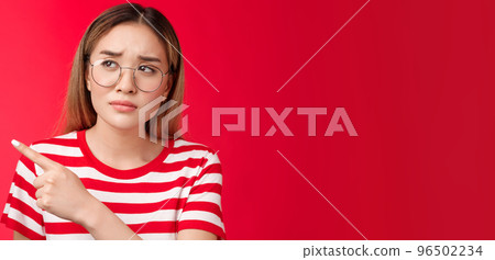 Disappointed doubtful frowning asian girl asking question reassure, looking pointing left displeased, unimpressed with strange place, dislike interiour sulking distressed, stand red background 96502234