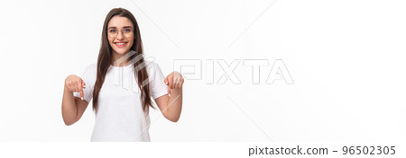 Click here. Portrait of brunette gorgeous young woman in glasses, pointing fingers down to show advertisement, smiling pleased, suggest visit link, recommend download or subscribe, white background 96502305
