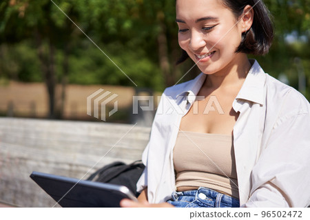 Portrait of young asian girl sitting in park with laptop, writing with digital pen or drawing, getting inspiration from outdoors Portrait of young asian girl sitting in park with laptop, writing with digital pen or drawing, getting inspiration from outdoors 96502472