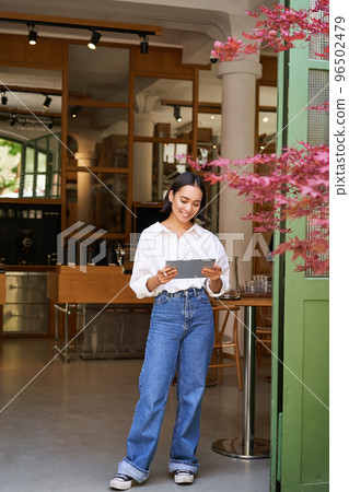 Young asian woman with tablet, standing in front of cafe entrance, inviting guests and customers, smiling broadly at camera Young asian woman with tablet, standing in front of cafe entrance, inviting guests and customers, smiling broadly at camera 96502479
