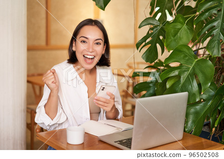 Joyful young asian woman winning, celebrating victory or achievement, receive good news on mobile phone, sitting with laptop and working Joyful young asian woman winning, celebrating victory or achievement, receive good news on mobile phone, sitting with laptop and working 96502508