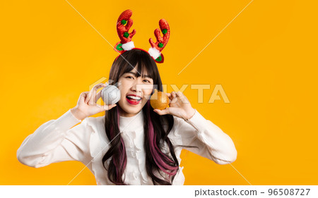 Pretty asian woman long hairstyle in white sweater wearing reindeer horns headband holding silver and gold glitter balls smiling and looking camera yellow background. Merry Christmas concept. 96508727