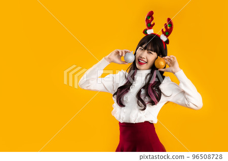 Asian woman long hair wear white and red cloth with reindeer horns headband Christmas theme costume holding gold and silver glitter balls looking camera yellow background. 96508728