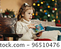 Portrait of a curly-haired little girl in a warm knitted sweater with a book in her hands against the background of a Christmas tree enjoying a cozy winter evening at home. Christmas, New Year 96510476